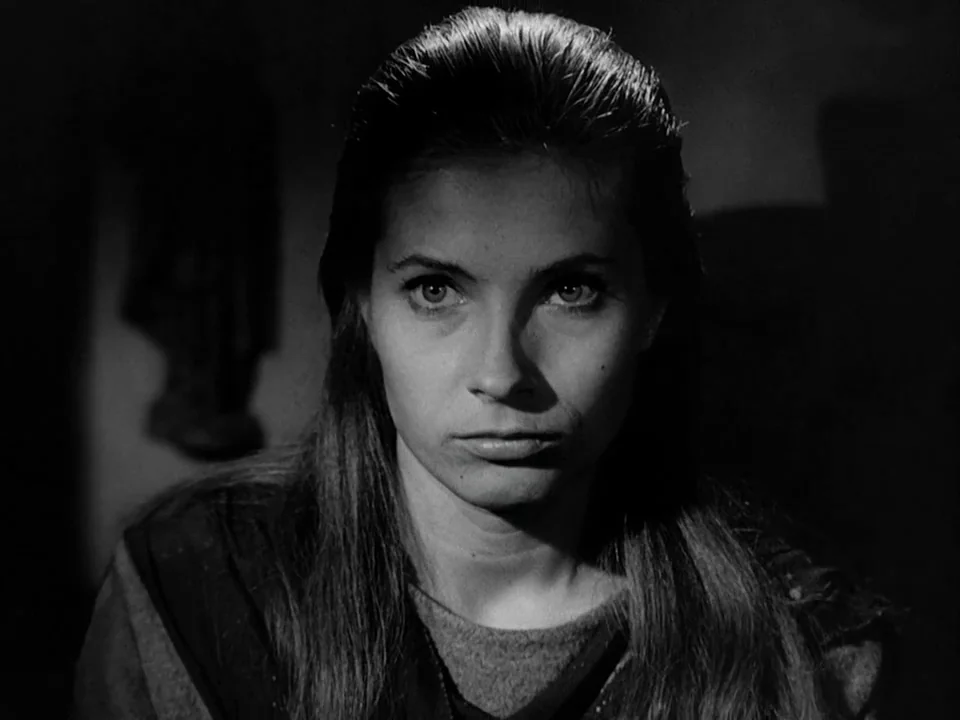 The Seventh Seal (1957) movie still: closeup — A dimly lit stone chamber with shadows obscuring the background. A woman looks directly ahead with a…