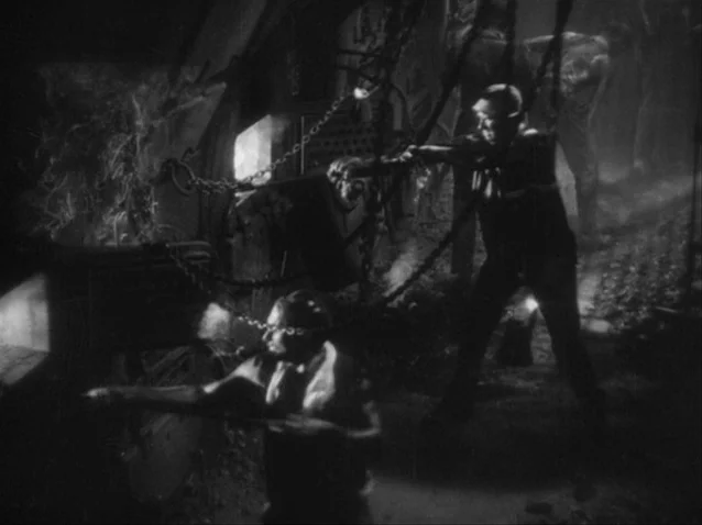 The Docks of New York (1928) movie still: medium — A dark industrial space filled with heavy chains and mechanical equipment. Two men perform labor…