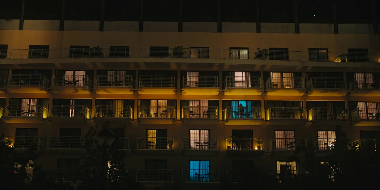 Sniper: G.R.I.T. (2023) movie still: wide — A multi-story hotel facade features rows of balconies and illuminated room windows at night. Most rooms…
