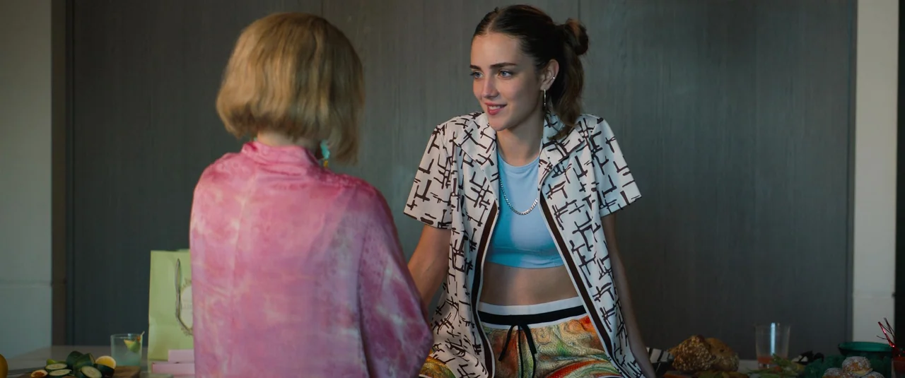 Do Revenge (2022) movie still: medium — A modern kitchen island surface holds various food items and a cocktail glass. A young woman sitting on the…