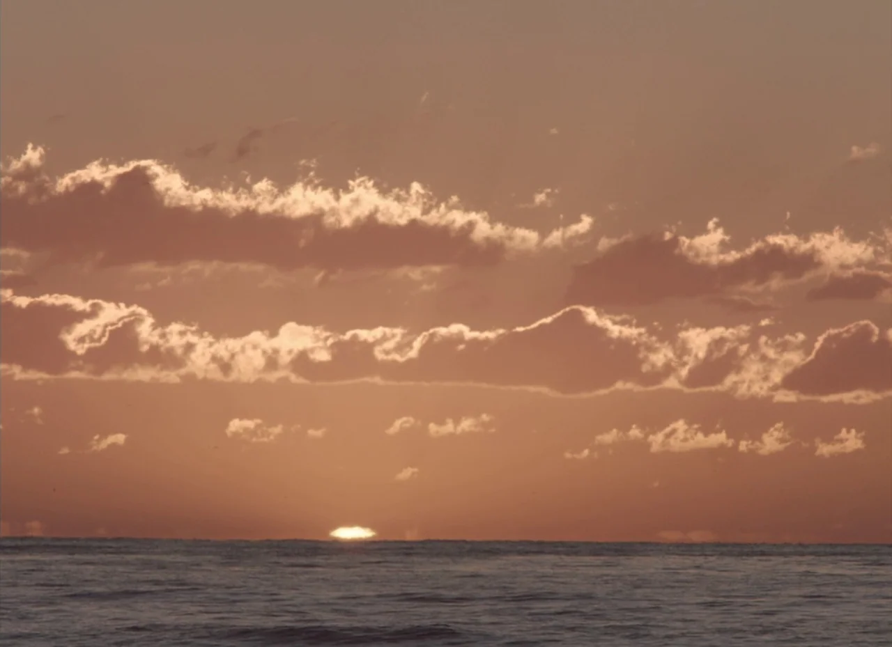 The Nightingale (2018) movie still: establishing — The sun sets over a calm ocean surface under a layer of scattered clouds. A thin sliver of the…