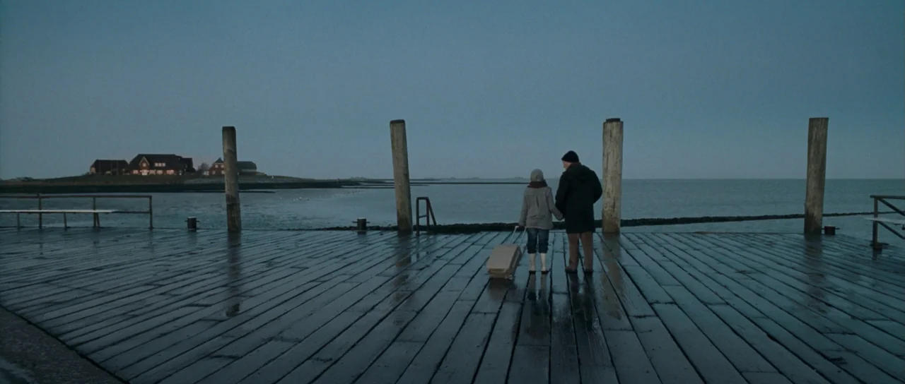 Womb (2010) movie still: wide — A long wooden pier extends over a calm, grey sea under an overcast sky. A child and an elderly man stand together on…