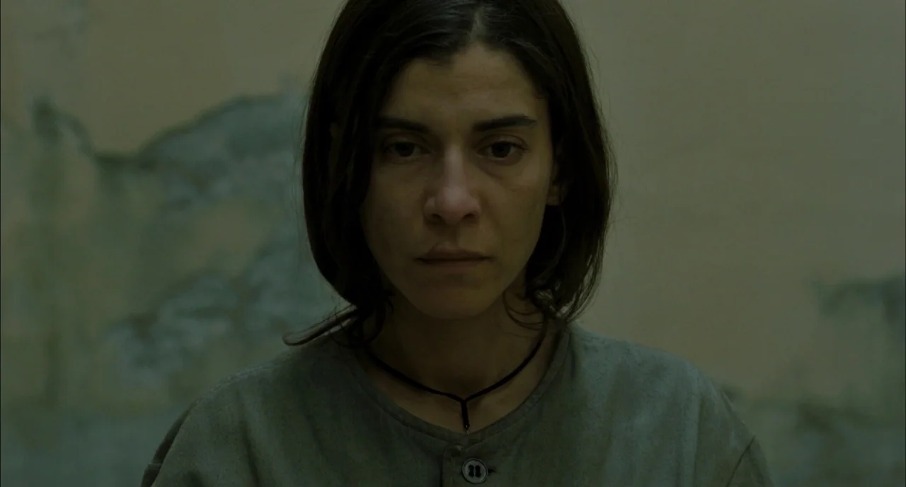 Incendies movie still: closeup — A woman stands in front of a wall with peeling, weathered paint textures. The subject maintains a direct, steady…