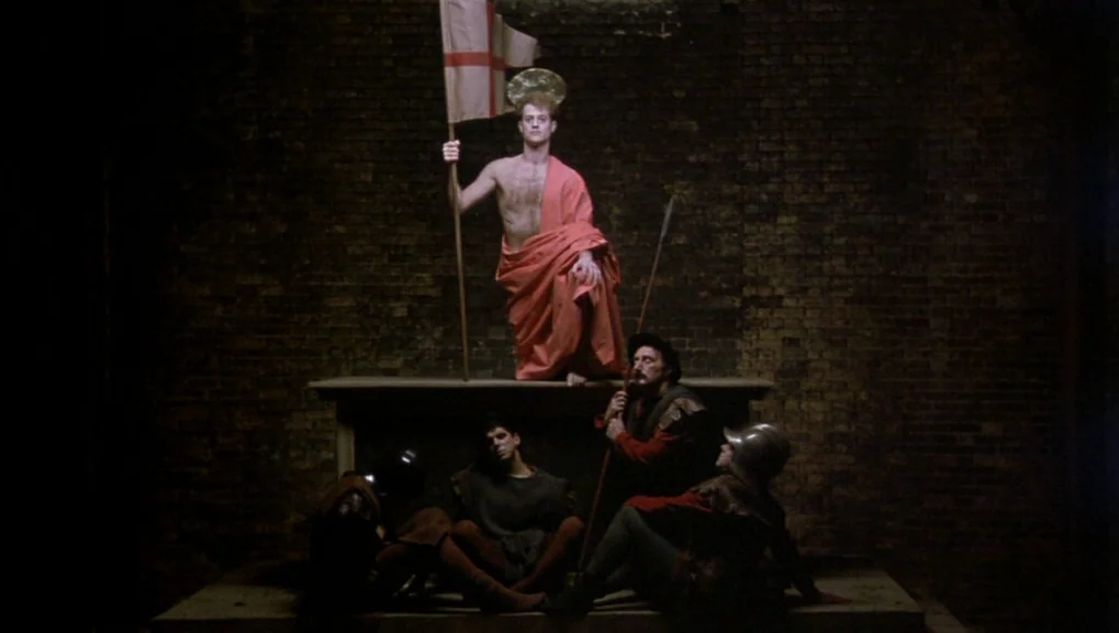 War Requiem (1989) movie still: wide — A group of people sits around a stone pedestal in front of dark brickwork. The central figure stands atop the…