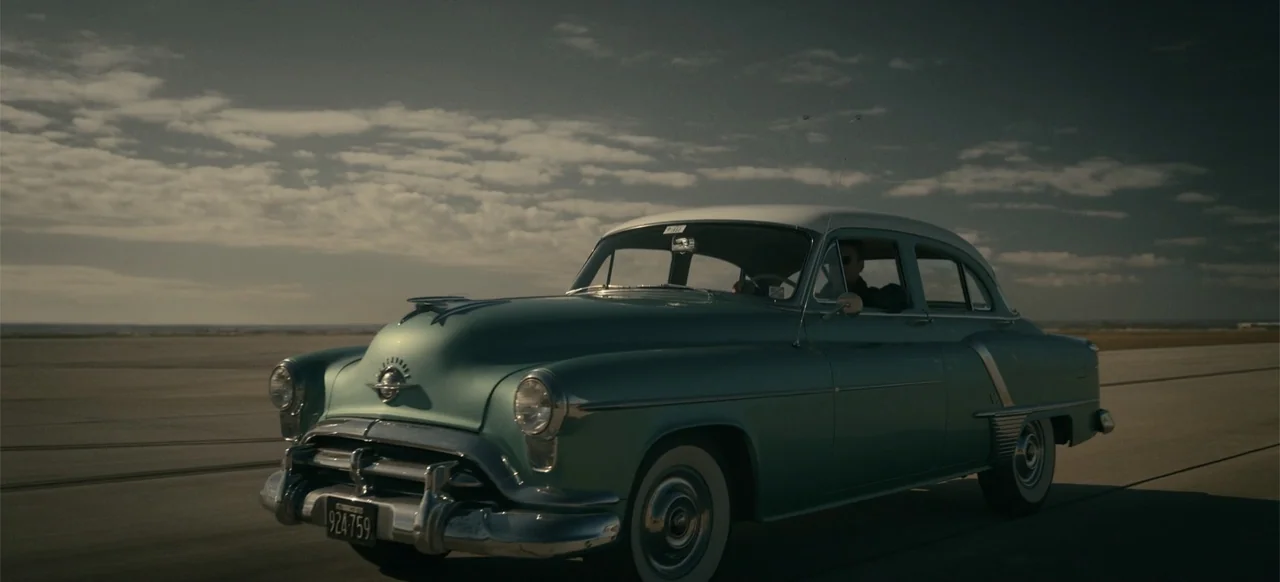Devotion (2022) movie still: wide — A light blue 1950s sedan drives across a vast, flat concrete runway during the day. The driver steers the…