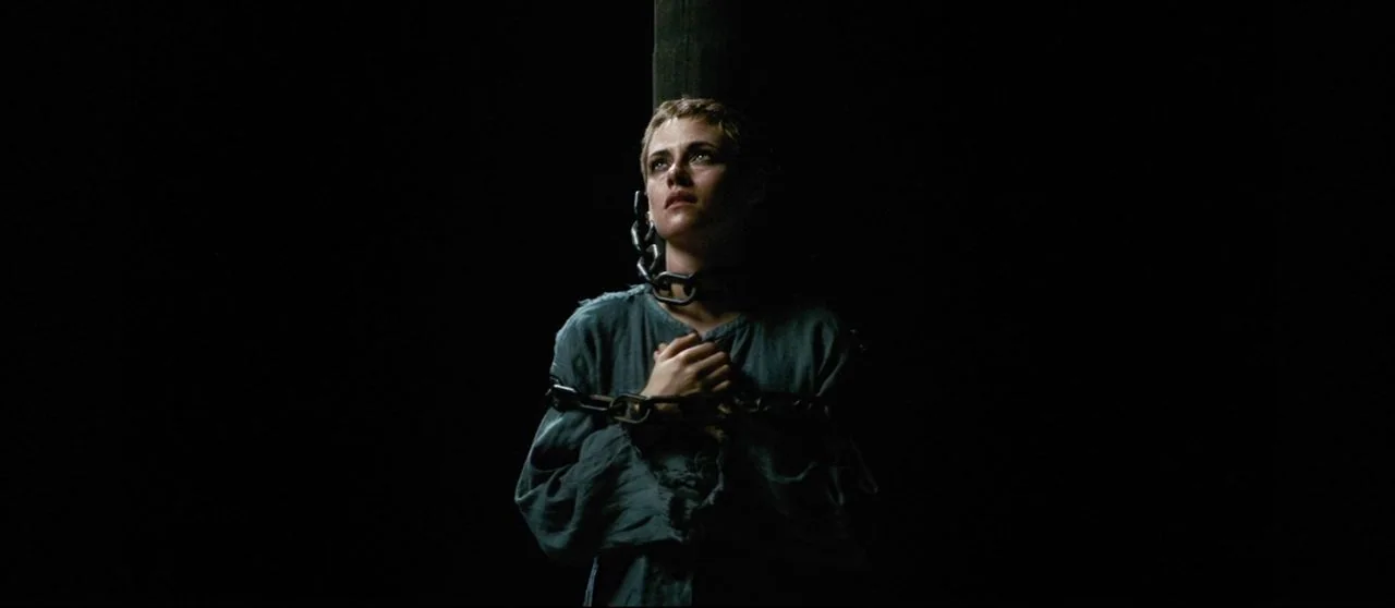 Seberg (2019) movie still: medium — A woman stands tied to a vertical wooden post in total darkness. She is chained by her neck and wrists against…