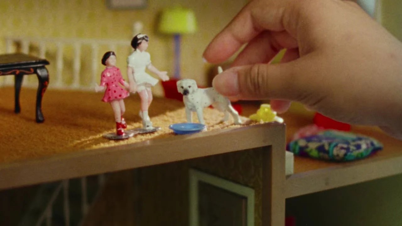 Shirkers (2018) movie still: closeup — A miniature wooden dollhouse room contains tiny plastic figurines of girls and a dog. A large hand enters the…