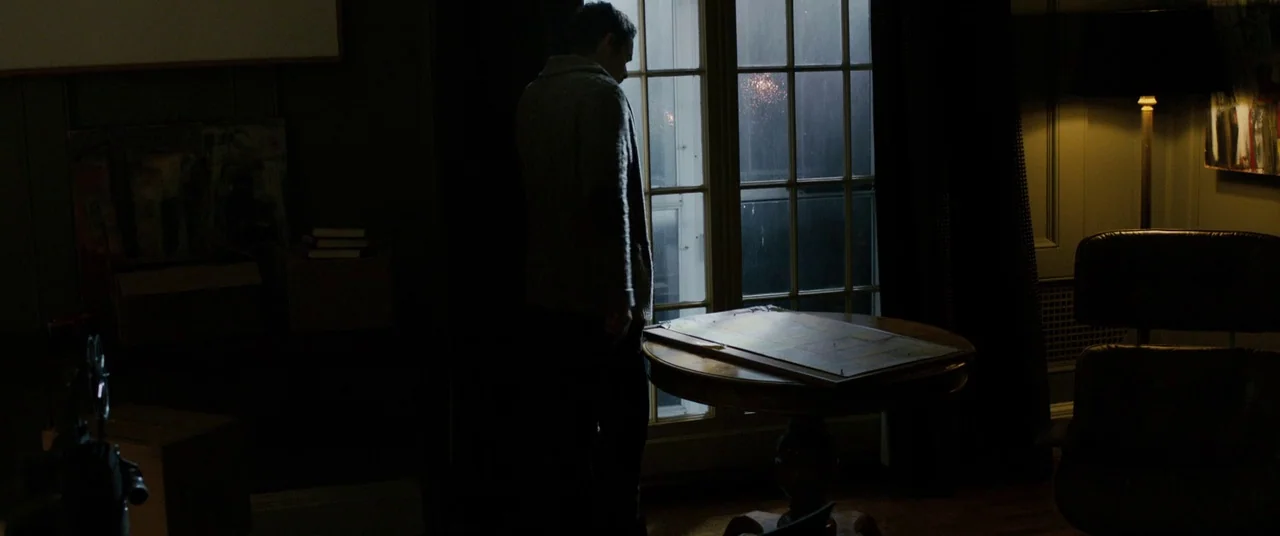 Sinister (2012) movie still: medium — A dark study features a single floor lamp and a large window at night. A man stands silently while observing a…