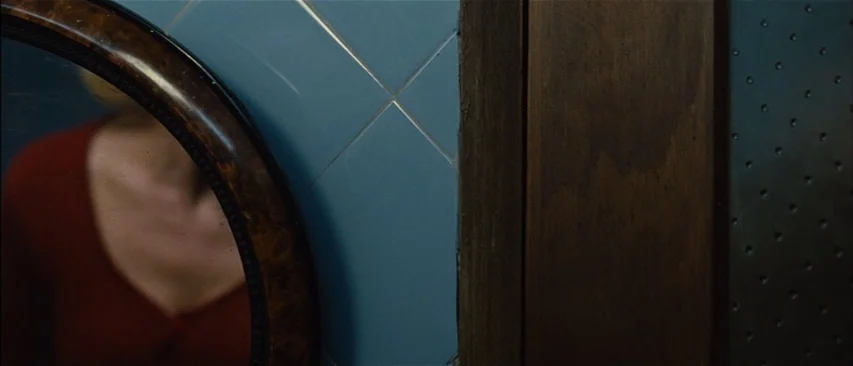 The Headless Woman (2008) movie still: closeup — A close-up shot focuses on a circular wooden mirror frame against blue tiled walls. A blurred…