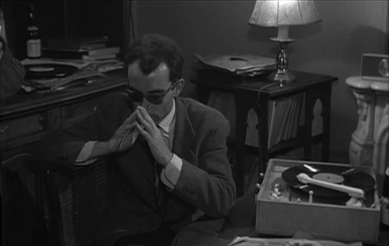 Le Signe du Lion (1962) movie still: medium — A living room interior featuring a desk, a record player, and a table lamp. A man sits at the table…