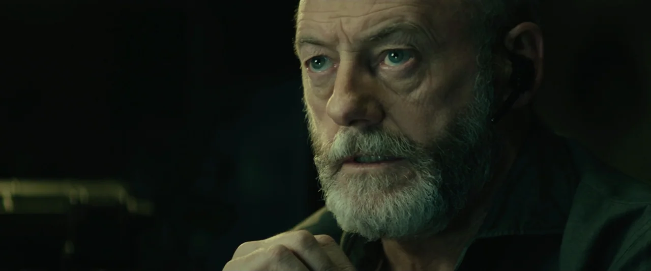 The Vault (2021) movie still: closeup — A dark office environment is dimly lit by a nearby light source. An older man with a gray beard watches…