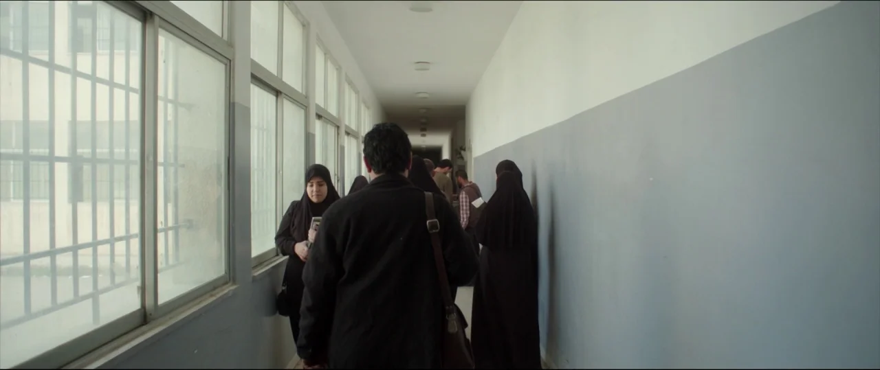 Under The Shadow (2016) movie still: wide — A long, institutional school hallway with large windows and a grey and white wall. Several people in…