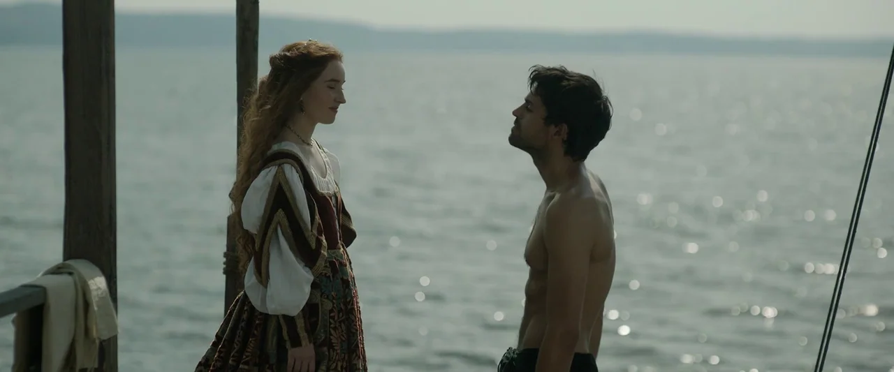 Rosaline (2022) movie still: medium — A wooden pier extends over a calm, reflective body of water. A woman in a historic-style gown stands facing a…