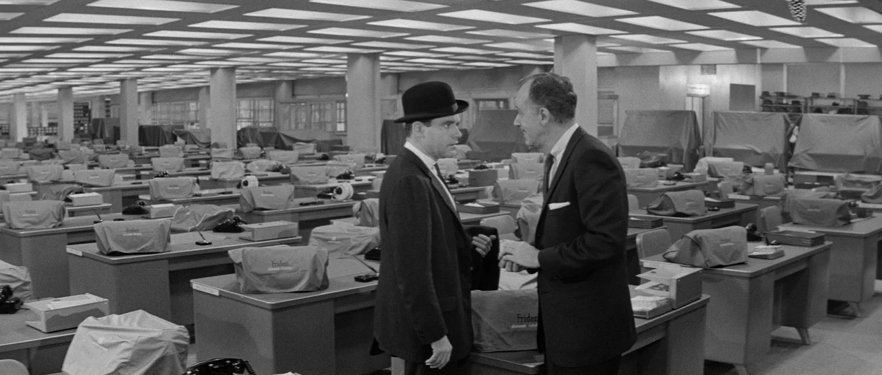 The Apartment (1960) movie still: medium — A vast, empty-looking office space is filled with rows of desks and covered machines. Two men in suits…