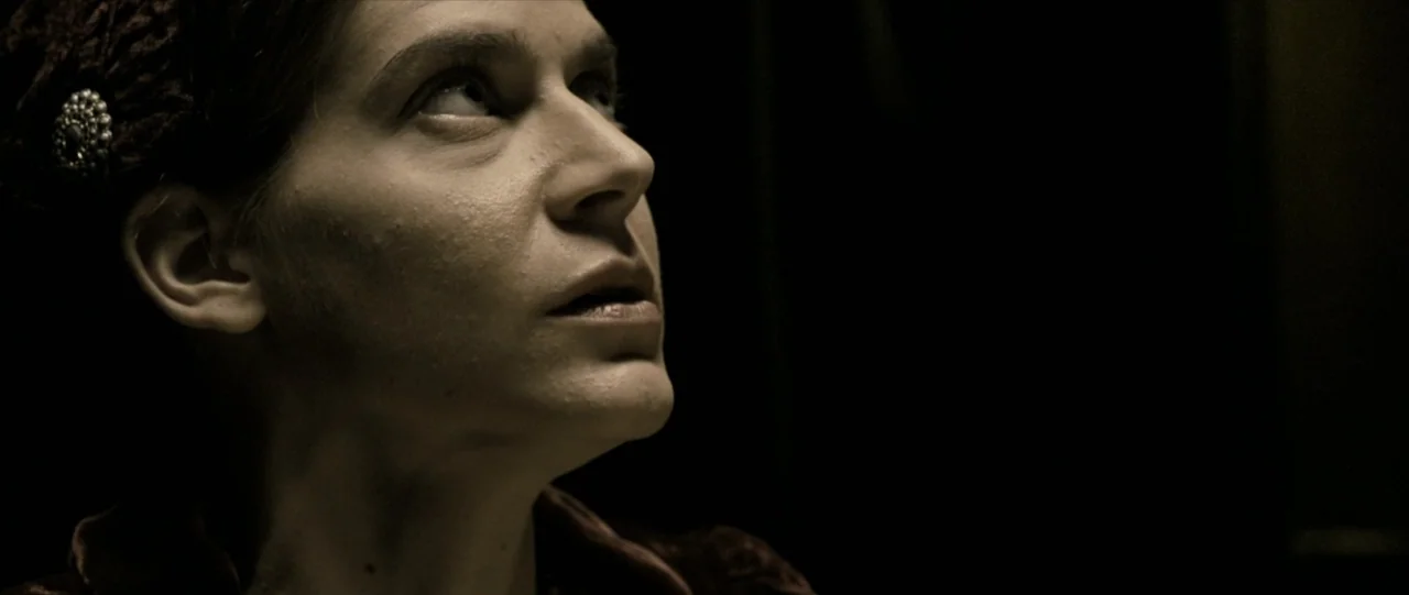 The Haunting in Connecticut (2009) movie still: closeup — A dark interior setting with a woman visible against a black background. She gazes upwards…