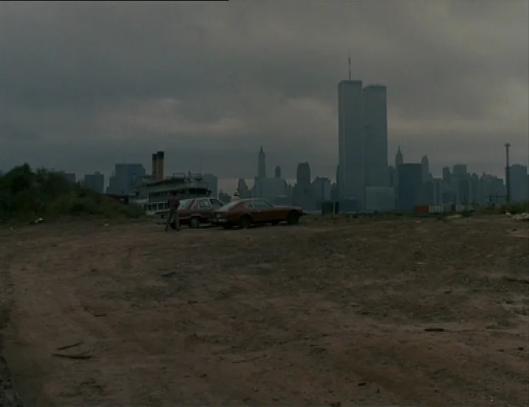 The Brother From Another Planet (1984) movie still: establishing — A vast, barren dirt lot overlooks a river with a distant city skyline. Two…