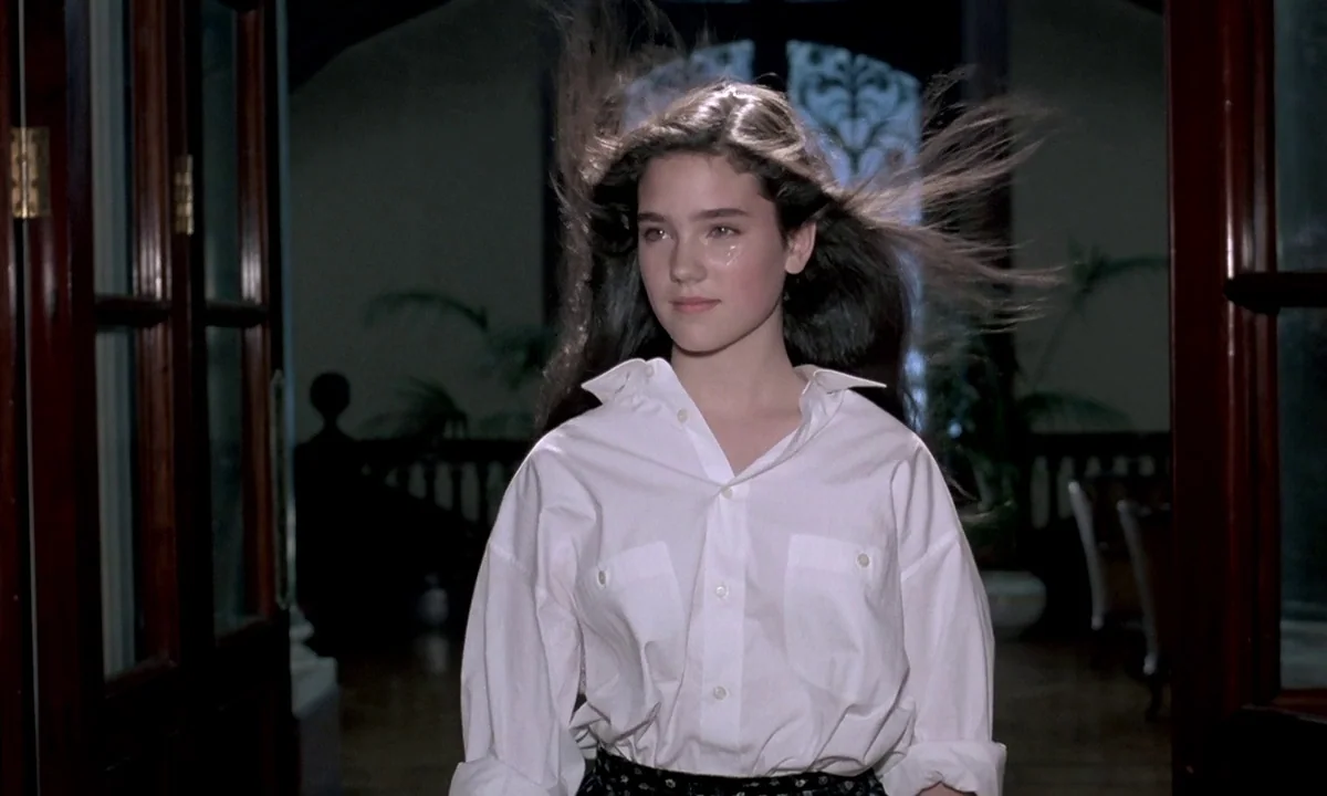 Phenomena (1985) movie still: medium — A spacious interior hallway with dark wood trim and large glass doors. A teenage girl walks through the open…