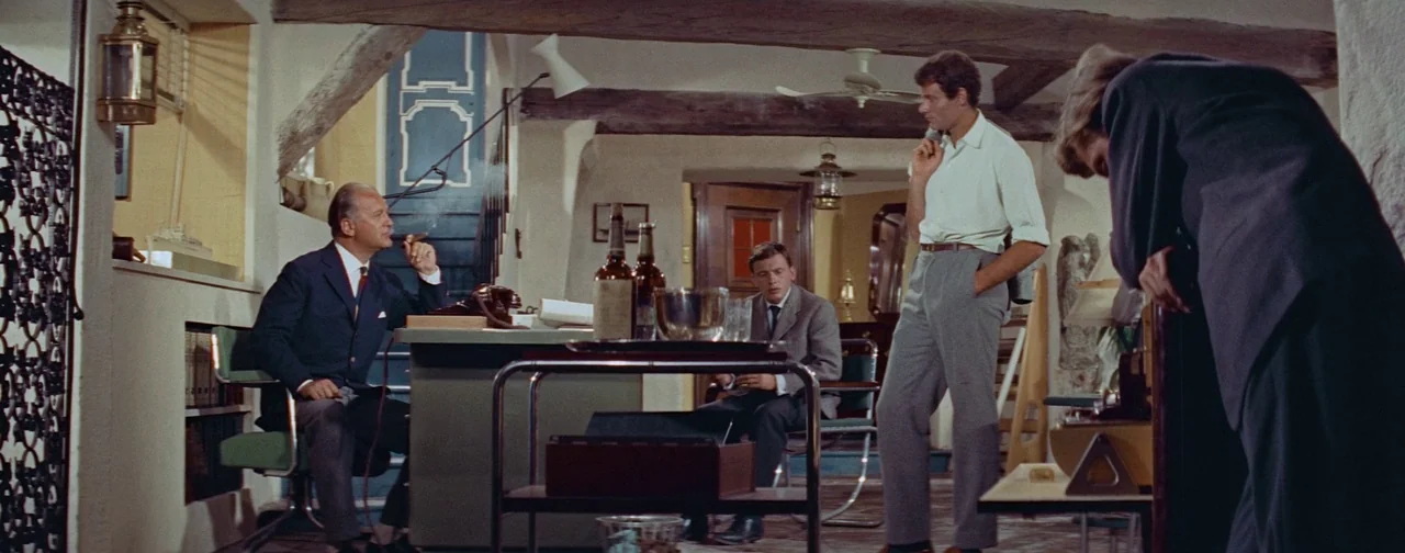 And God Created Woman (1956) movie still: wide — A vintage 1960s study features wooden ceiling beams, a desk, and mid-century furniture pieces. Four…