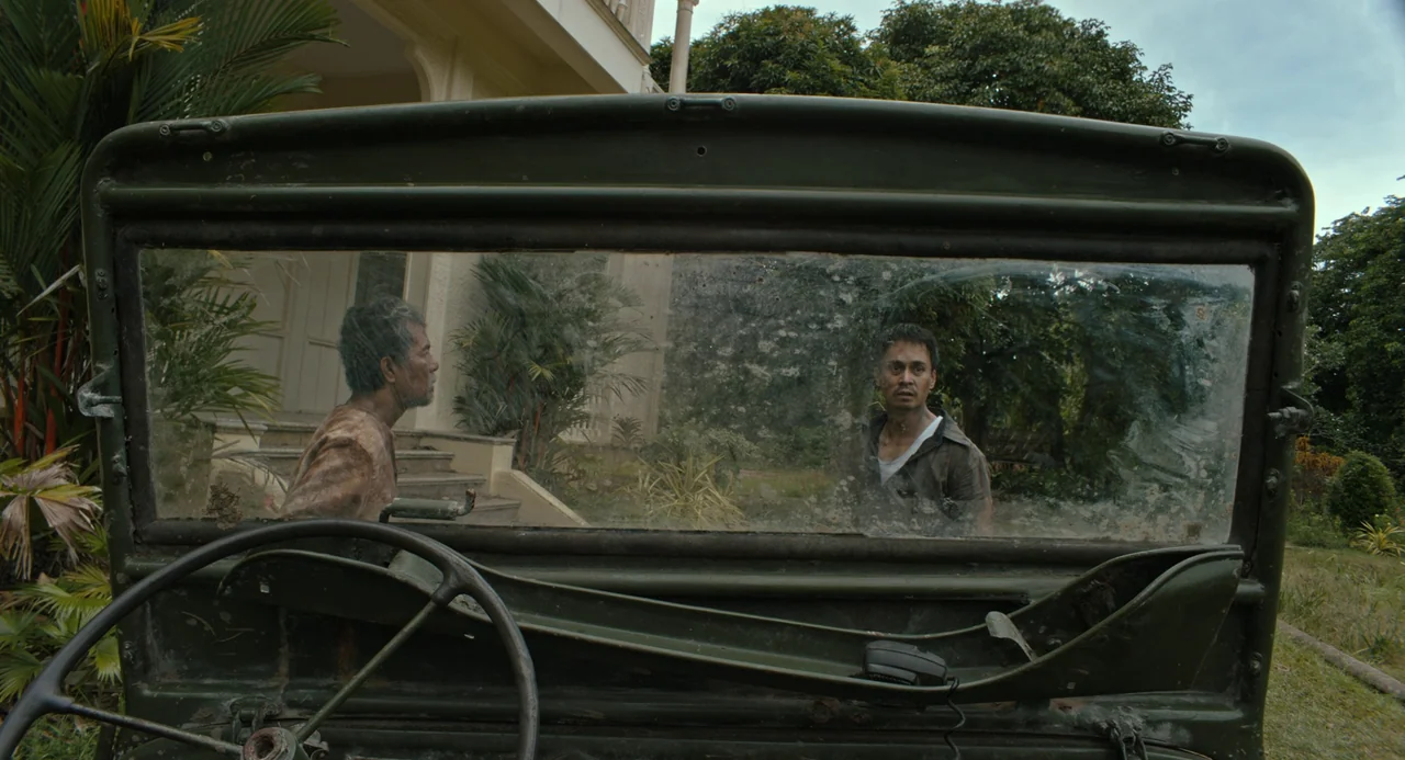 In My Mother’s Skin (2023) movie still: medium — Two men face each other through the dusty windshield of a vintage military-style jeep. A large…