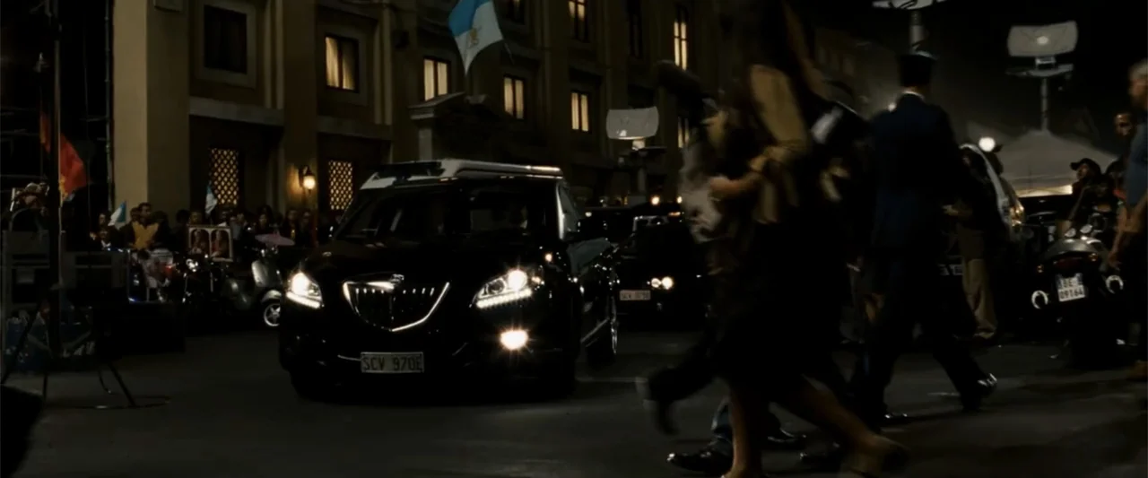 Angels & Demons (2009) movie still: medium — A black sedan drives down a dimly lit city street at night. Pedestrians walk past the car while other…