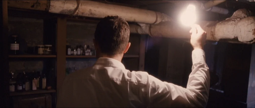 The Killer Inside Me (2010) movie still: medium — A dark, cramped basement with exposed plumbing pipes running along the low ceiling. A man holds a…