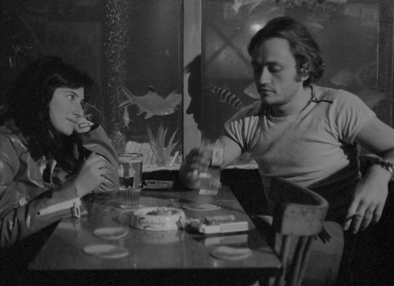 Je Tu Il Elle (1974) movie still: medium — A dark bar booth features a large aquarium built into the back wall. A young couple sits across from each…