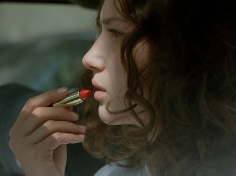 Hail Mary (1985) movie still: closeup — The interior of a vehicle on a bright day. A young woman applies bright red lipstick to her lips.