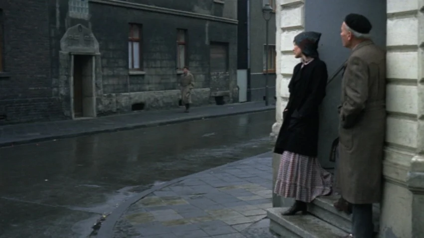 Tenderness of the Wolves (1973) movie still: fullbody — A wet concrete urban street with dark stone buildings and a solitary pedestrian nearby. An…