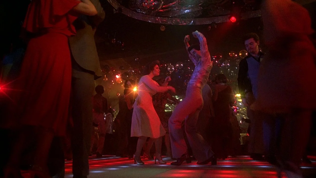 Saturday Night Fever (1977) movie still: fullbody — A crowded discotheque with a glowing illuminated dance floor and overhead mirror balls. A man in…