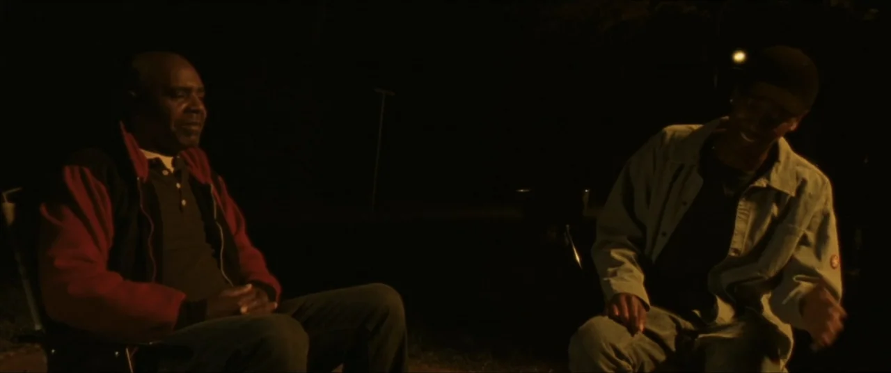 Sermon on the Mount (2019) movie still: medium — Two men sit outside at night in folding chairs illuminated by a nearby fire. The older man listens…