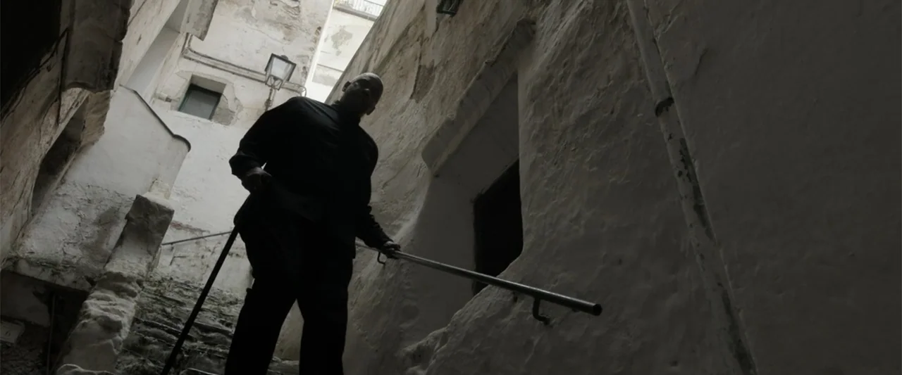 The Equalizer 3 (2023) movie still: fullbody — A narrow exterior stone staircase sits between two high, weathered white-painted walls. A man in dark…