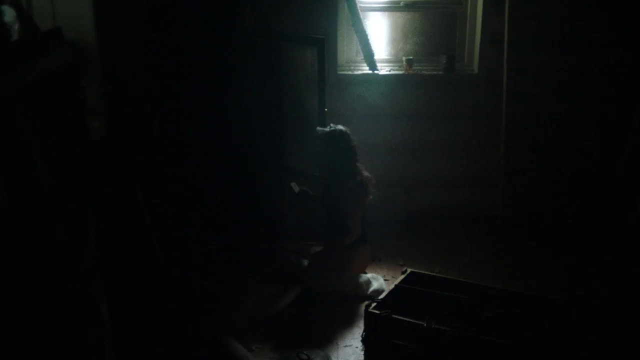 Snowbound (2017) movie still: wide — A dark room with sparse furnishings is illuminated by a single window. A woman kneels on the floor in profile,…