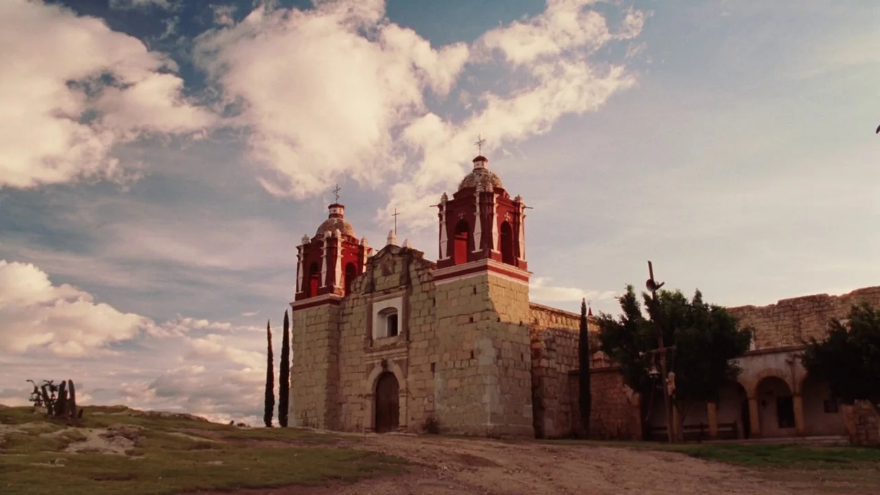 Nacho Libre (2006) movie still: establishing — A historic stone church with red towers sits on a dry rural landscape. The structure is flanked by…
