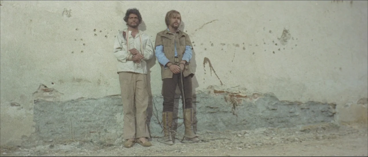 The Mercenary (1968) movie still: fullbody — A weathered plaster wall shows bullet impact pockmarks and crumbling stucco at the base. Two men stand…