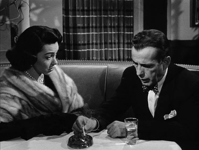 In a Lonely Place (1950) movie still: medium — A dimly lit restaurant booth features patterned curtains in the dark background. A man and a woman…