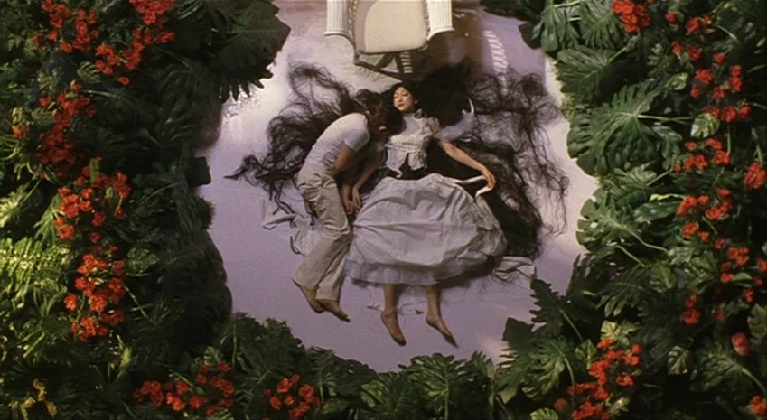 Rampo Noir (2005) movie still: wide — A white floor is surrounded by a dense border of green monstera leaves and red flowers. A man and a woman lie…