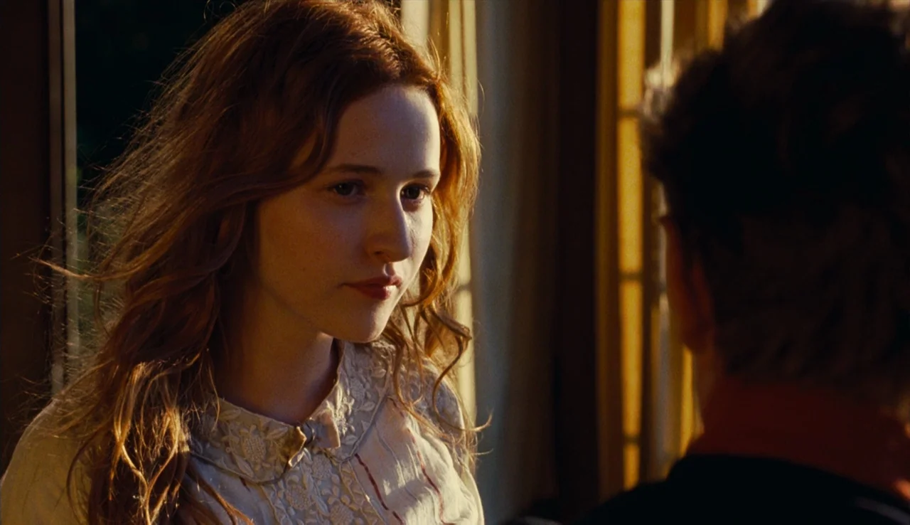 Renoir (2012) movie still: closeup — A dimly lit room features a woman standing near a window during sunset hours. She gazes toward a person…