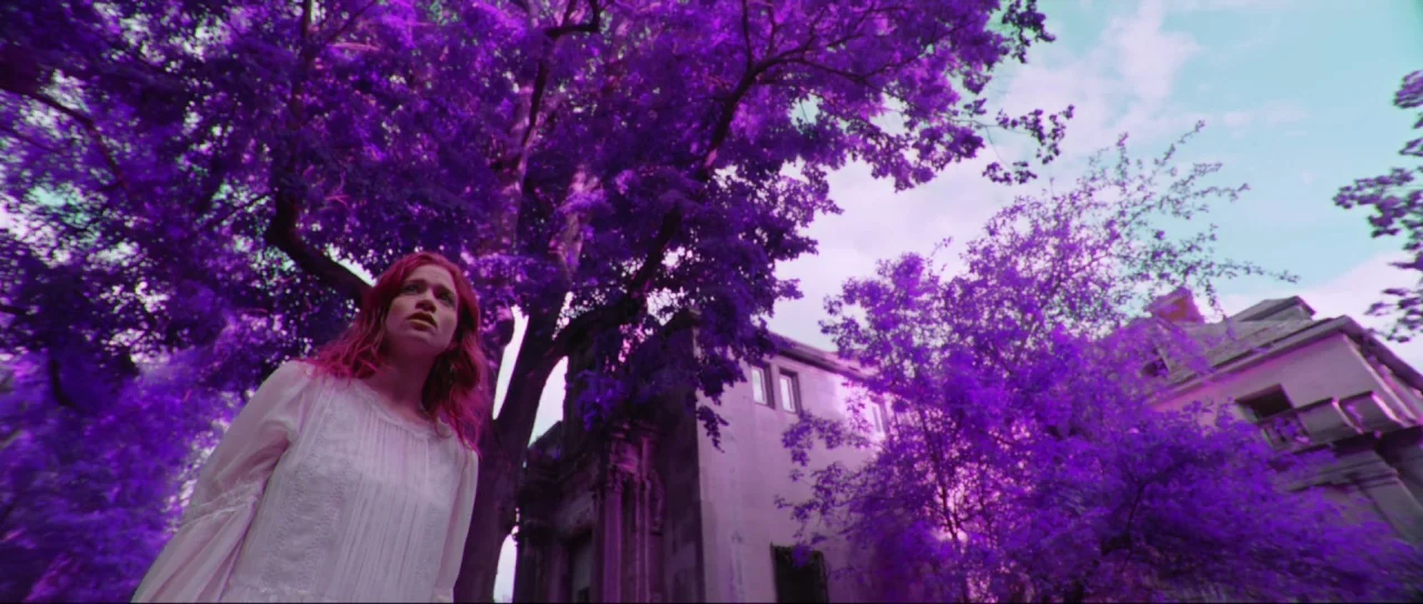 Braid (2018) movie still: medium — A woman stands in front of a stone building beneath large trees. The entire scene features an infrared effect…