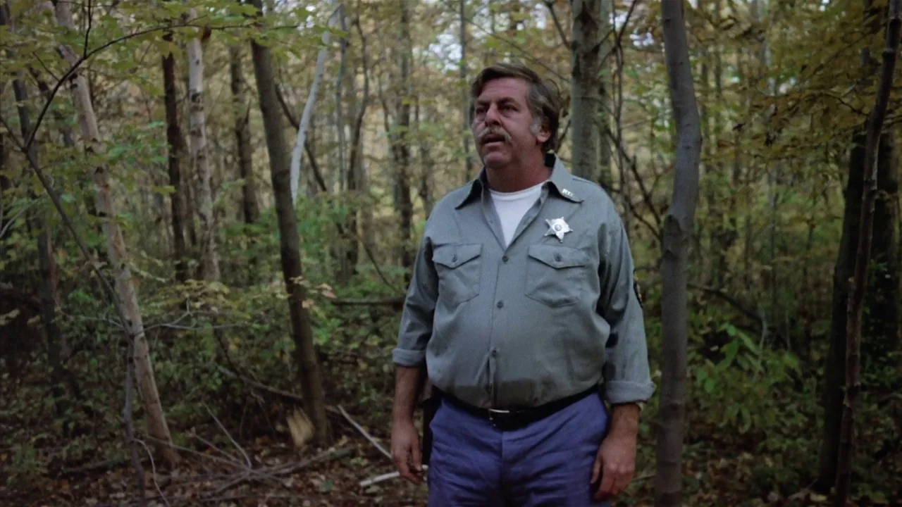 Friday the 13th Part 2 (1981) movie still: medium — A dense autumn woodland with thin, vertical tree trunks filling the background. A middle-aged…
