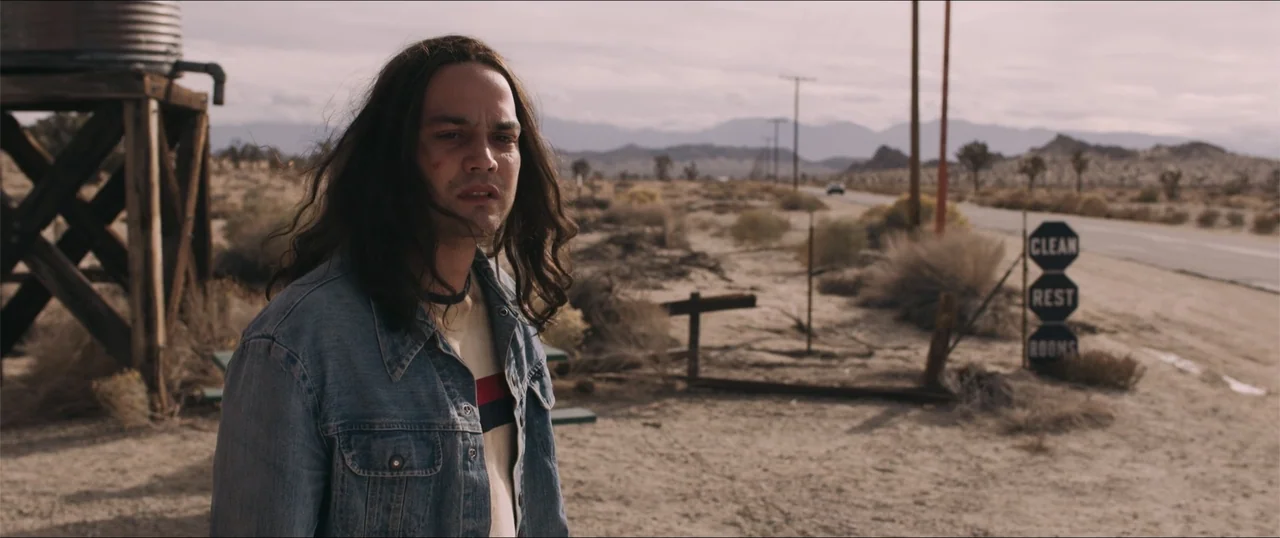 Woman of the Hour (2024) movie still: medium — A desolate desert roadside features a rusted water tank and signage for restrooms. A young man with…