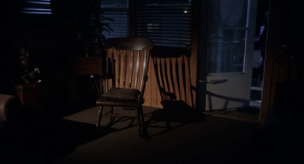 Happer’s Comet (2022) movie still: medium — A dimly lit room features wood paneling and closed window blinds at night. An empty rocking chair sits…