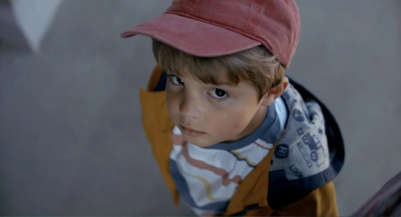 Living Out Loud (1998) movie still: closeup — A young boy stands outdoors on a grey paved surface. He is wearing a yellow jacket and red cap while…