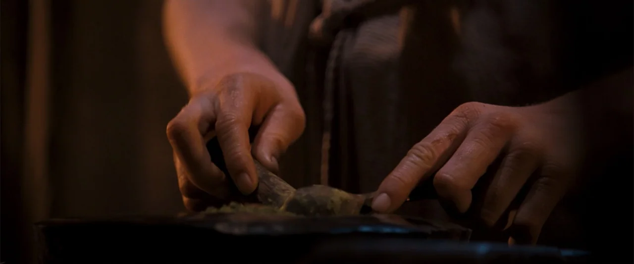 10,000 BC (2008) movie still: closeup — A dark interior workspace features hands working over a stone mortar and pestle. The person is grinding dry…