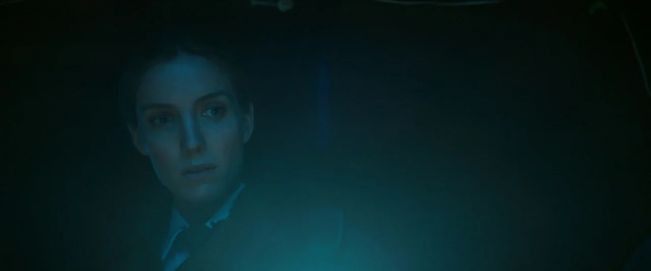 The Silencing (2020) movie still: closeup — A woman stands in a dark environment illuminated by a low-positioned light source. The subject looks…