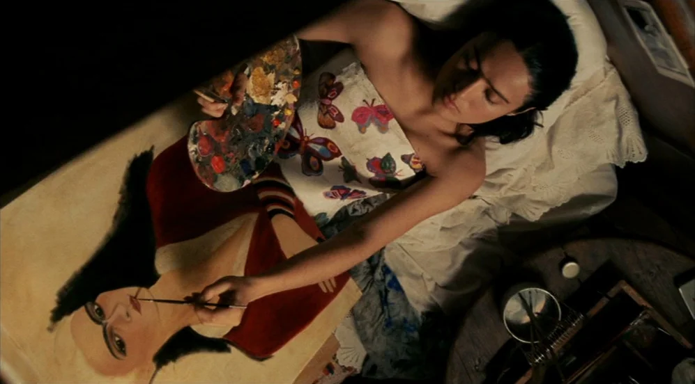 Frida (2002) movie still: medium — A woman lies on a bed, painting a self-portrait on a canvas propped before her. Her white top is covered with…