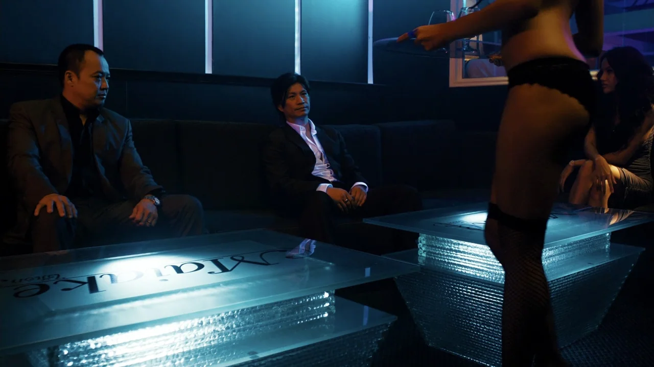 Zero Tolerance (2015) movie still: medium — A dark nightclub booth features two men seated on a sofa near illuminated tables. A woman in black…