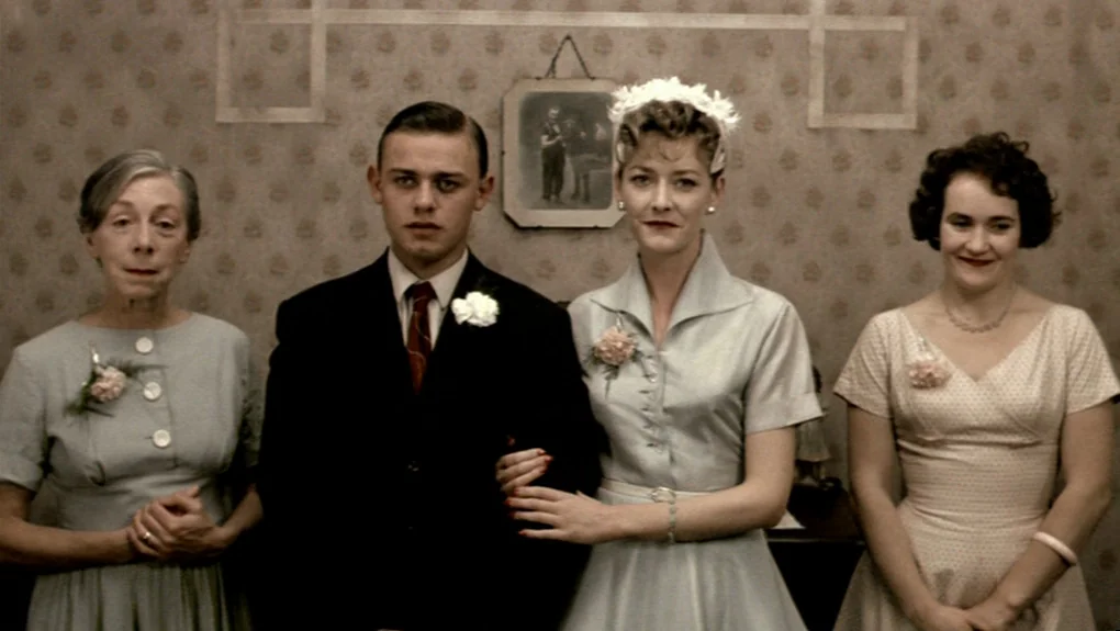 Distant Voices, Still Lives (1988) movie still: fullbody — A 1950s living room features vintage floral wallpaper with a framed photograph hanging…