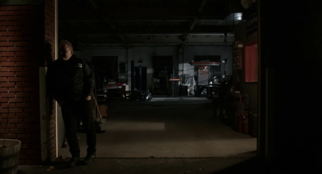 Happer’s Comet (2022) movie still: wide — A dark auto repair shop interior features a brick entrance pillar and concrete floors. A man wearing a…