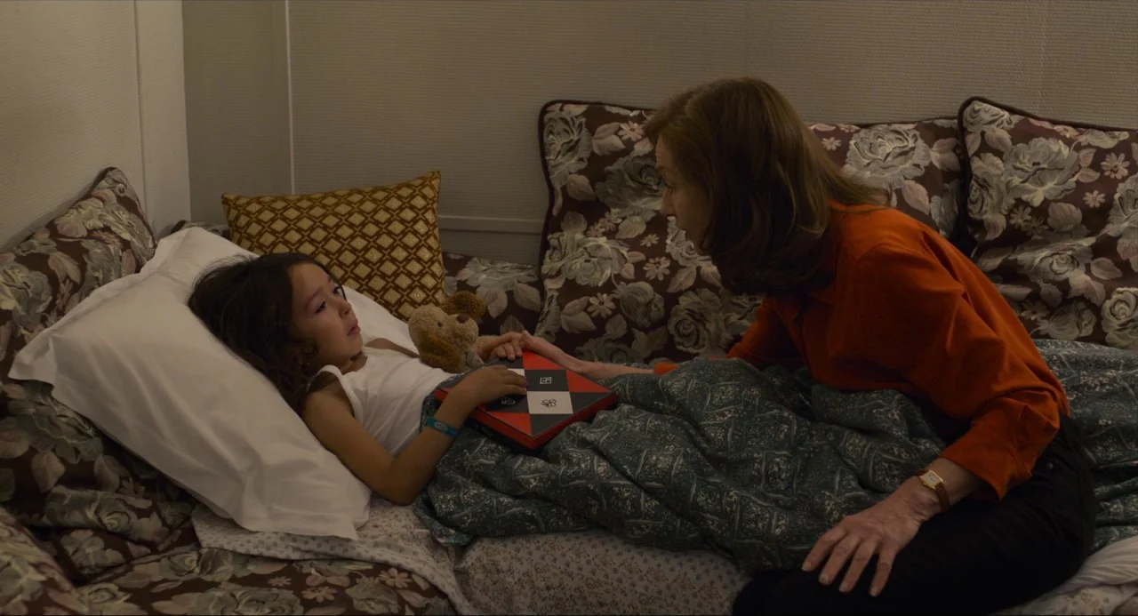 Happy End (2017) movie still: medium — A child rests in a bed with floral patterned linens and pillows. A woman leans toward the child while holding…