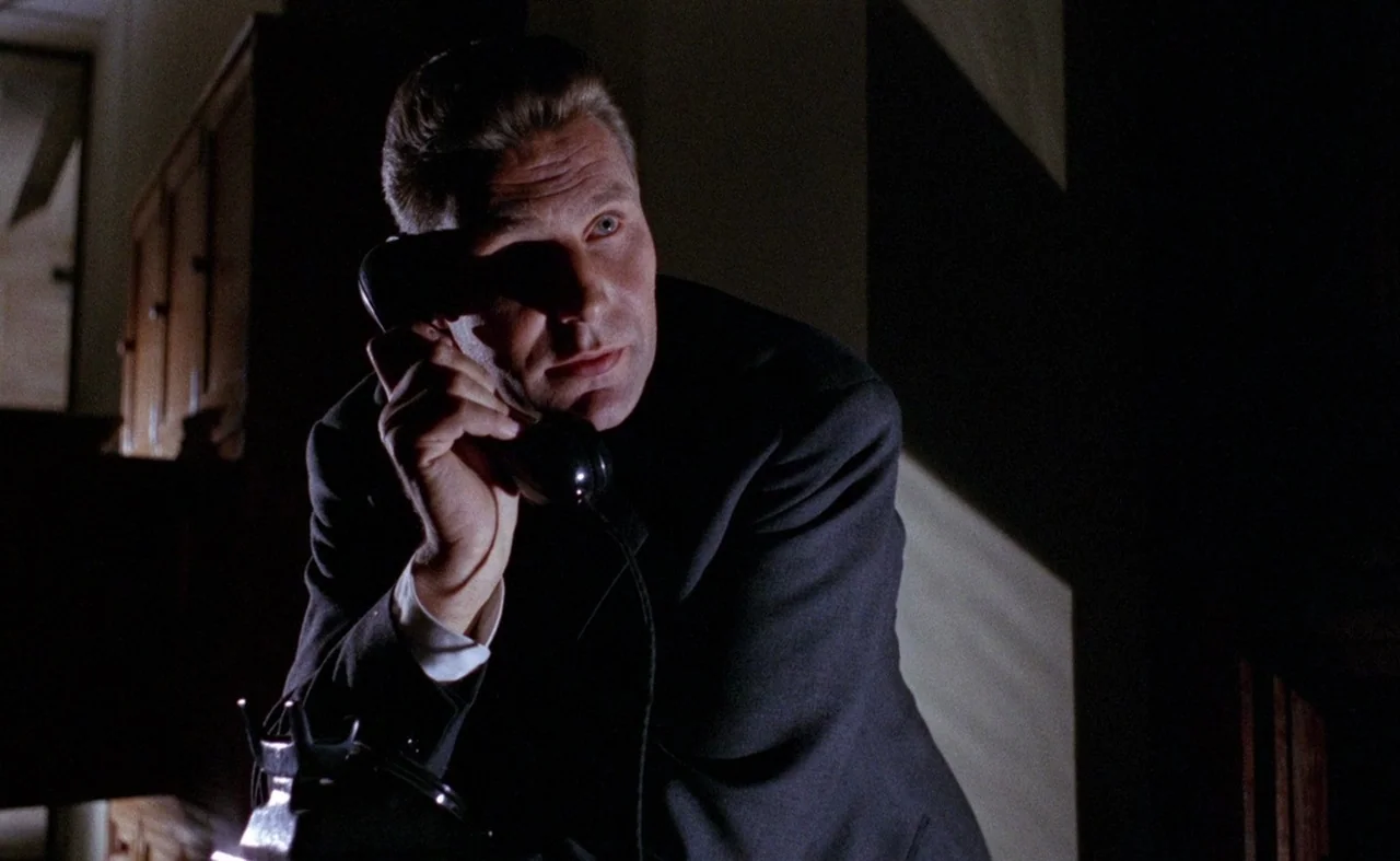 Blood And Black Lace (1964) movie still: medium — A dark, dimly lit hallway serves as the setting for this quiet moment. A man in a suit listens…